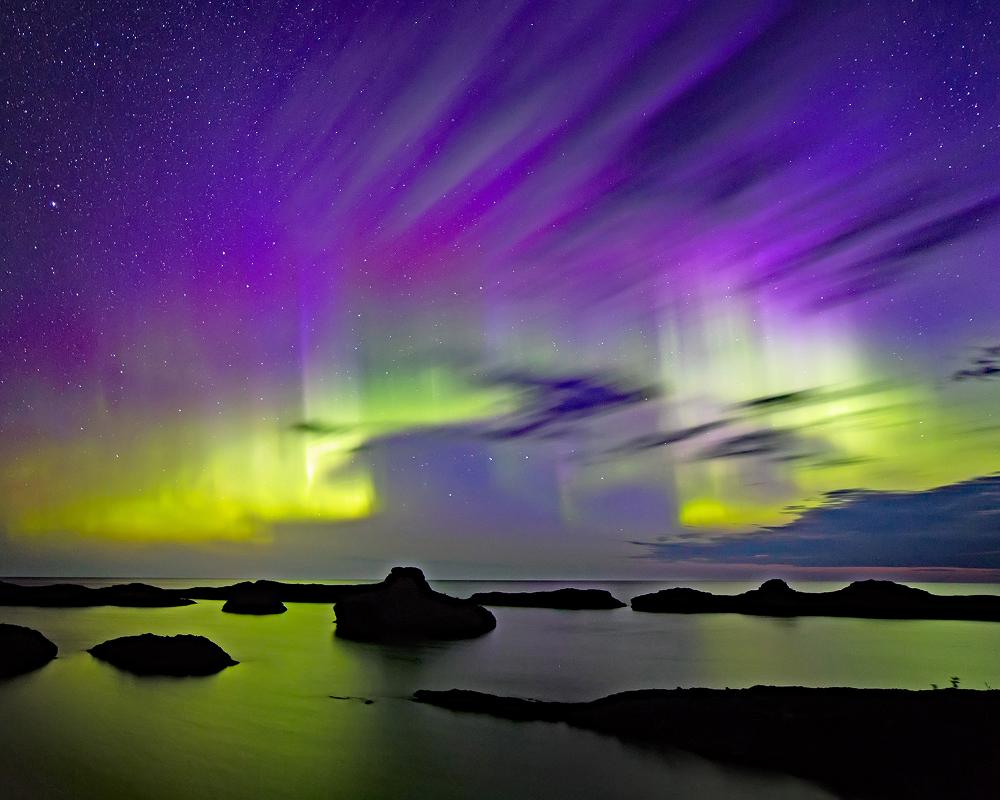the aurora borealis lights up a night sky over a series of rocks in a body of water